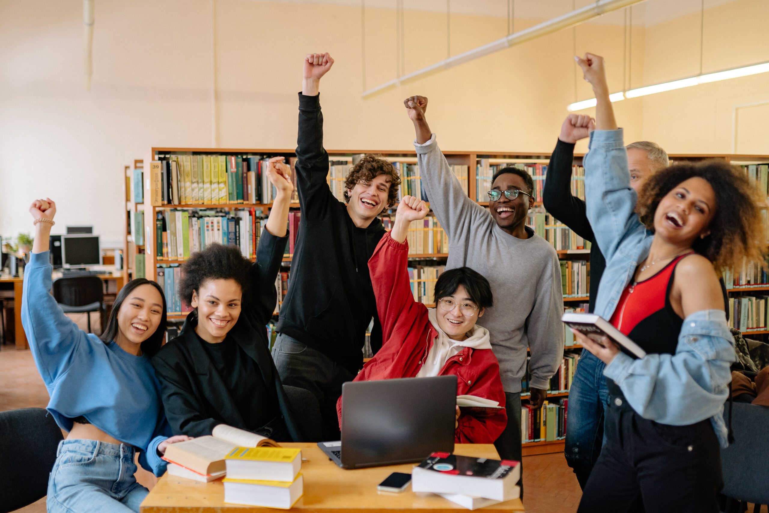 Grupa studentów w bibliotece podnosząca pięści do góry w geście zwycięstwa z uśmiechami na twarzach