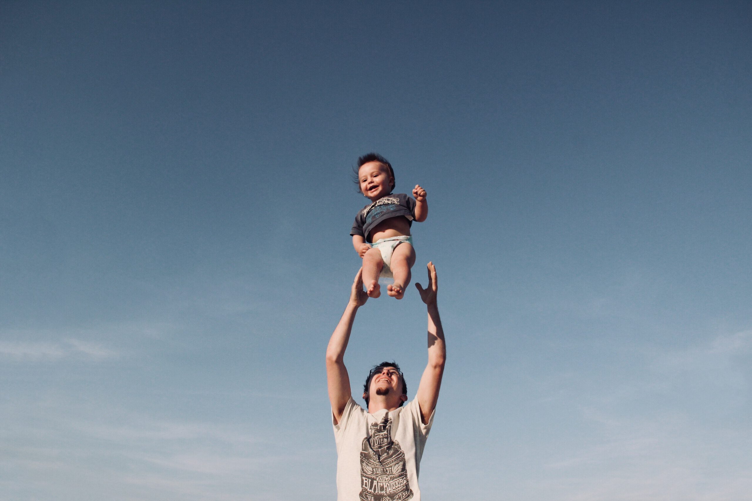 Ojciec podrzucający dziecko do góry na tle niebieskiego nieba